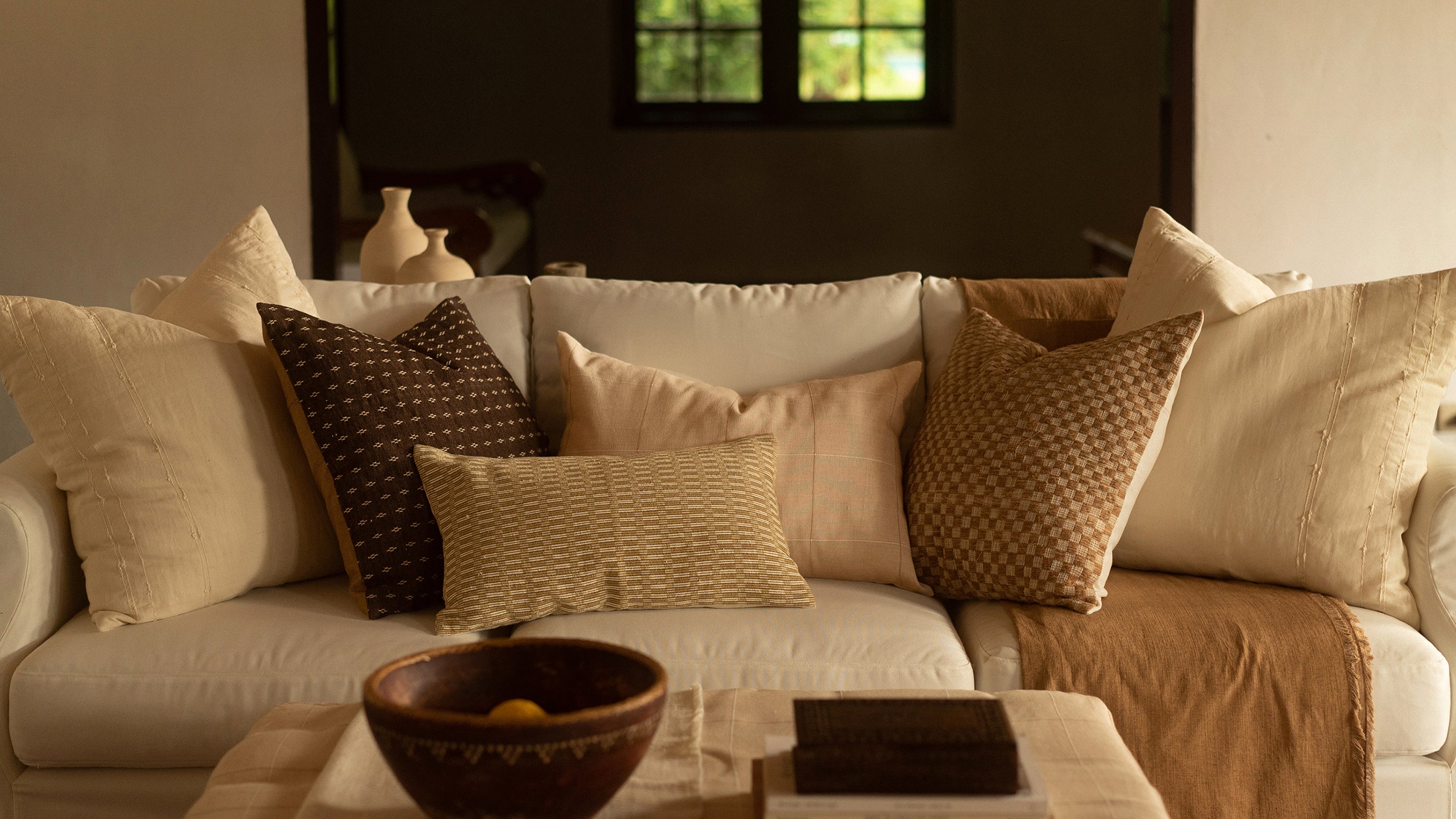 A cottage-style living room filled with earthy palette handwoven pillows and antique objects. It is furnished with a white sofa and twin armchairs in front of a fireplace. 